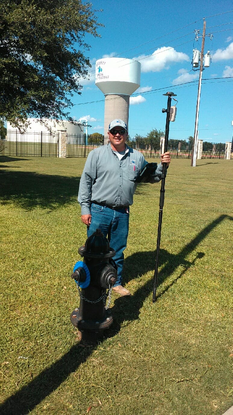 Benjamin Rucks stands outside the Rockett SUD office with one of the utility’s water towers in the background. He is holding his mobile GIS solution for field data collection, using ArcGIS Field Maps, an Eos Arrow Gold RTK receiver, and iPad